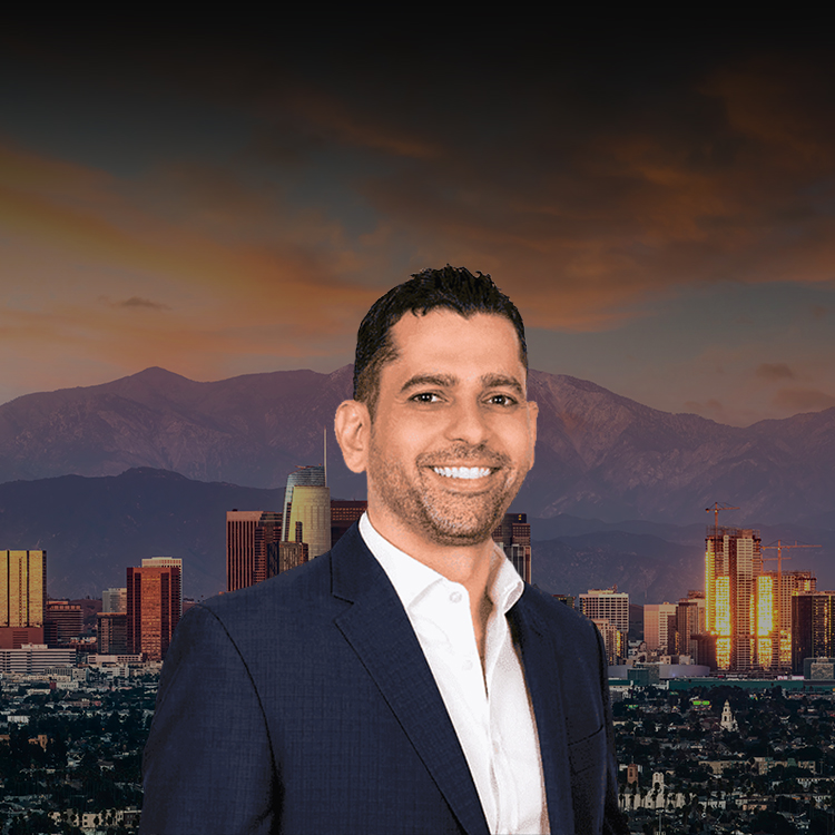 Photo of Steve Baghoomian, smiling, wearing a dark suit and a white shirt, stands in front of a blurred cityscape with skyscrapers at sunset.