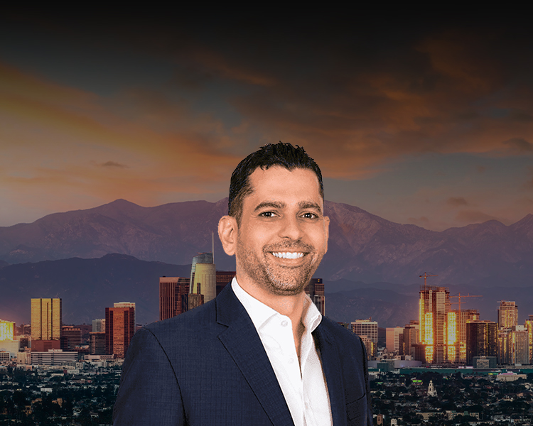Photo of Steve Baghoomian, smiling, wearing a dark suit and a white shirt, stands in front of a blurred cityscape with skyscrapers at sunset.